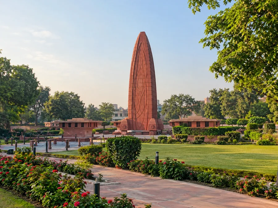 Das hohe, flammenförmige Denkmal aus rotem Sandstein in Jallianwala Bagh, zwischen Rasenflächen und Blumenbeeten